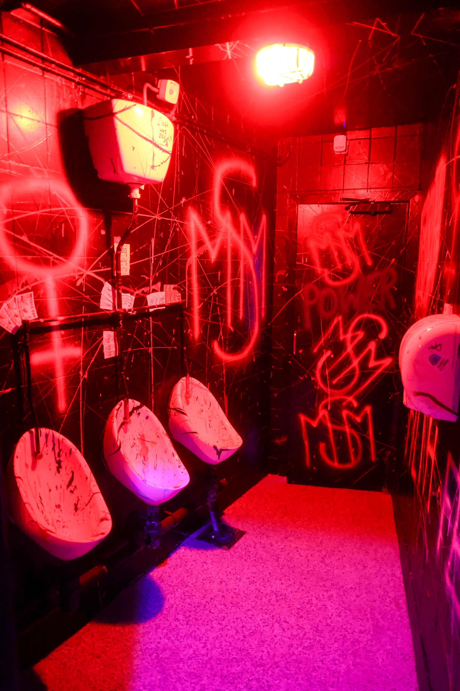 The Boys room in red neon light, with three urinals and MSM graffiti on the walls