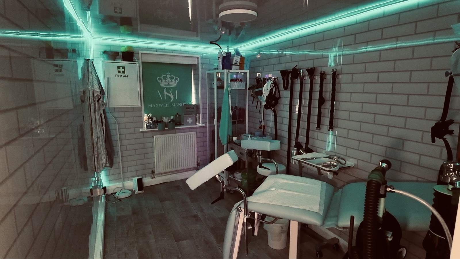 The Clinic: a white tiled room under teal neon with an examination chair, instrument trolley and first aid cabinet