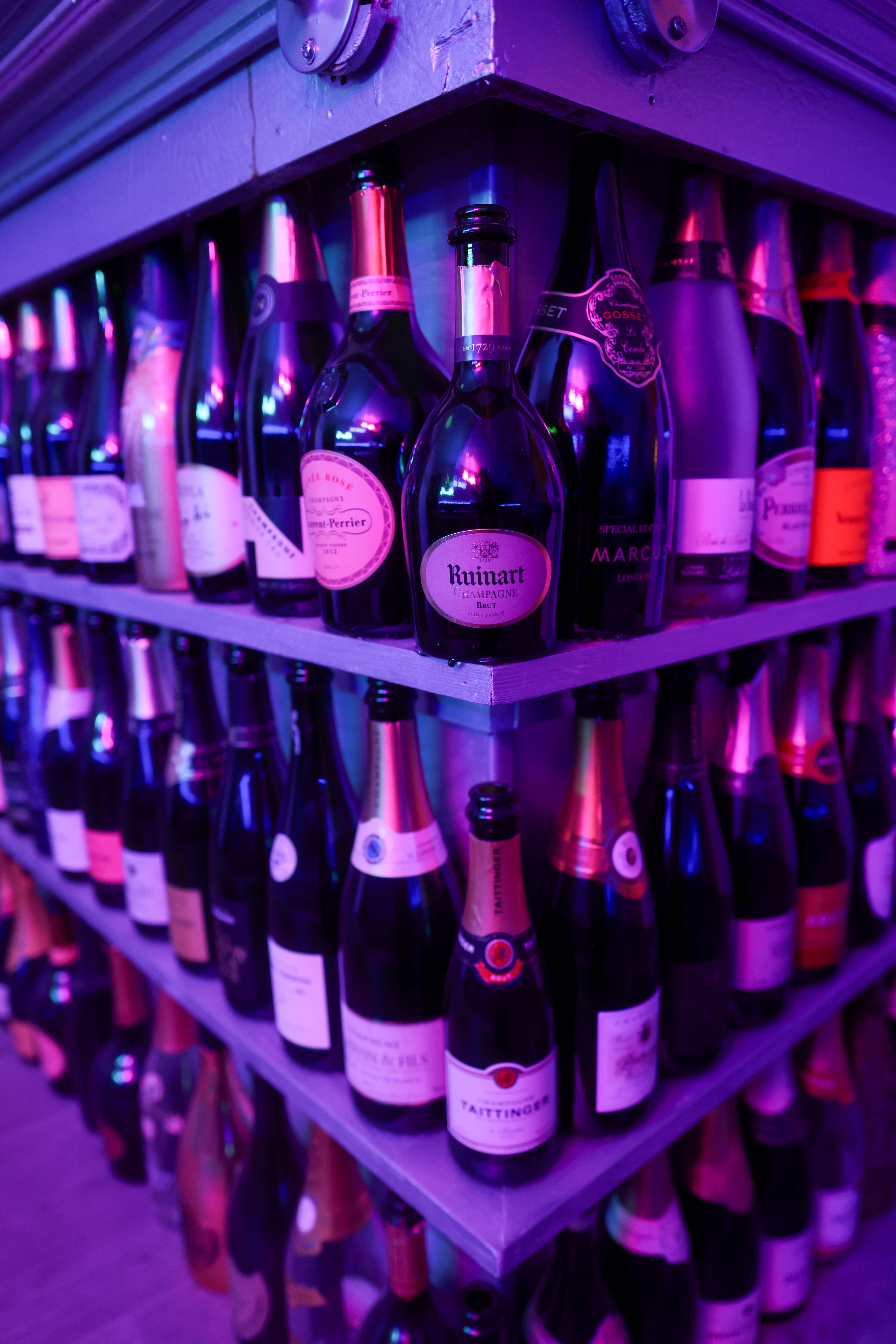 Shelves of champagne bottles including Ruinart and Taittinger, lit violet