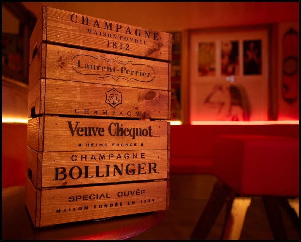 Stacked wooden champagne crates branded Bollinger, Veuve Clicquot and Laurent-Perrier in the Club’s red-lit lounge