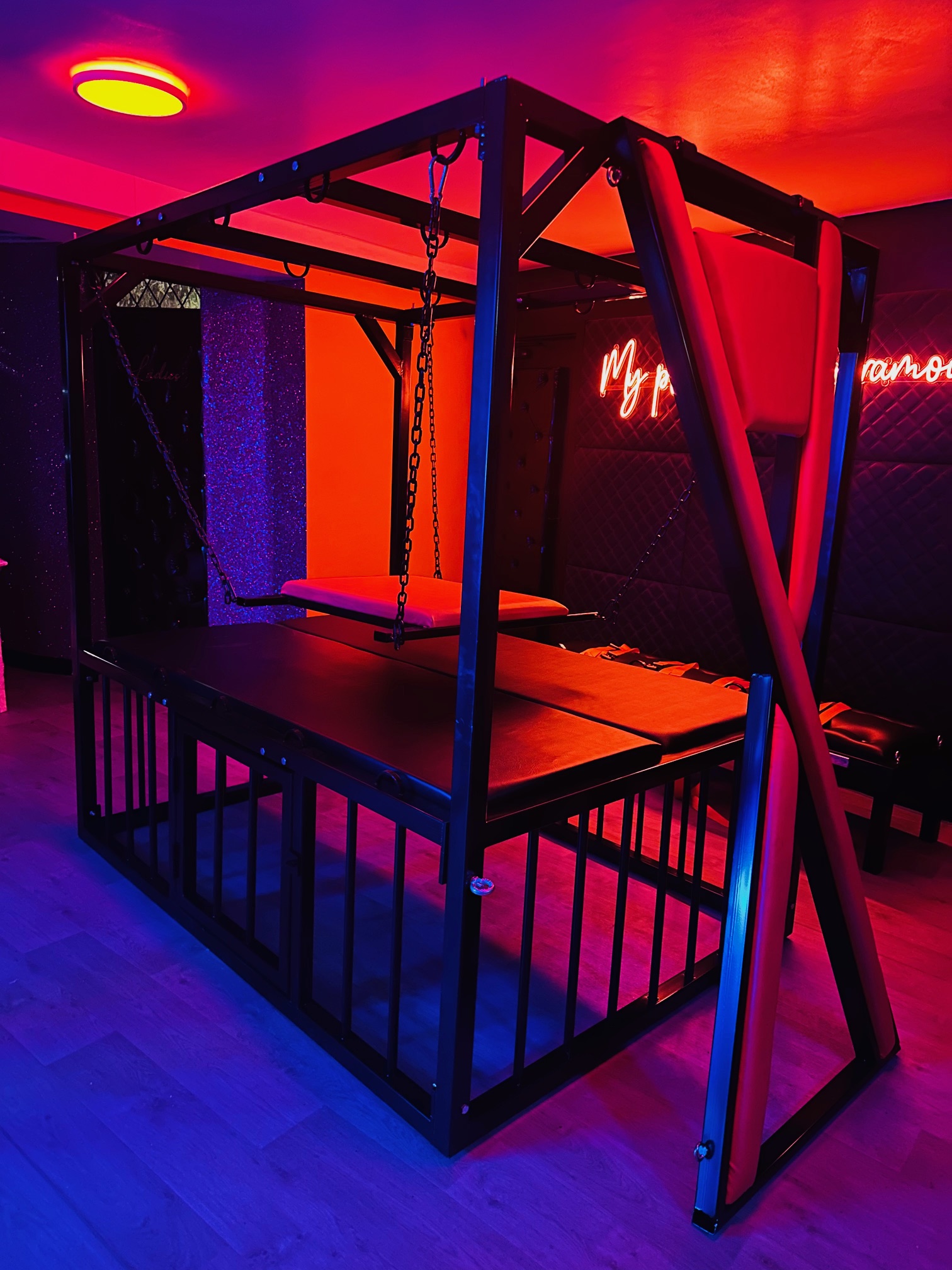 A black steel play frame with a chain-hung swing and padded table, lit orange and violet beneath a neon sign